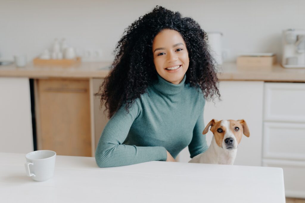Curly\-haired woman in turtleneck sits at kitchen table, enjoys tea, plays with Jack Russell terrier\. Joyful moment with beloved pet\.